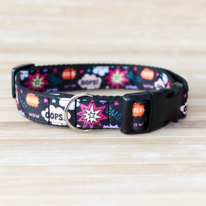 May include: Black dog collar with a comic book style design. The collar features colorful speech bubbles with words like "OMG!" and "OOPS!" along with starburst graphics. It has a black plastic buckle and a silver metal D-ring.