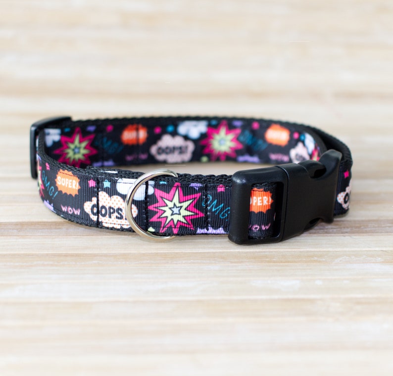 May include: Black dog collar with a comic book style design. The collar features colorful speech bubbles with words like "OMG!" and "OOPS!" along with starburst graphics. It has a black plastic buckle and a silver metal D-ring.