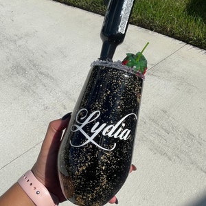 May include: Black and gold glitter tumbler with the name "Lydia" written in white cursive. The tumbler has a straw and a red strawberry garnish.