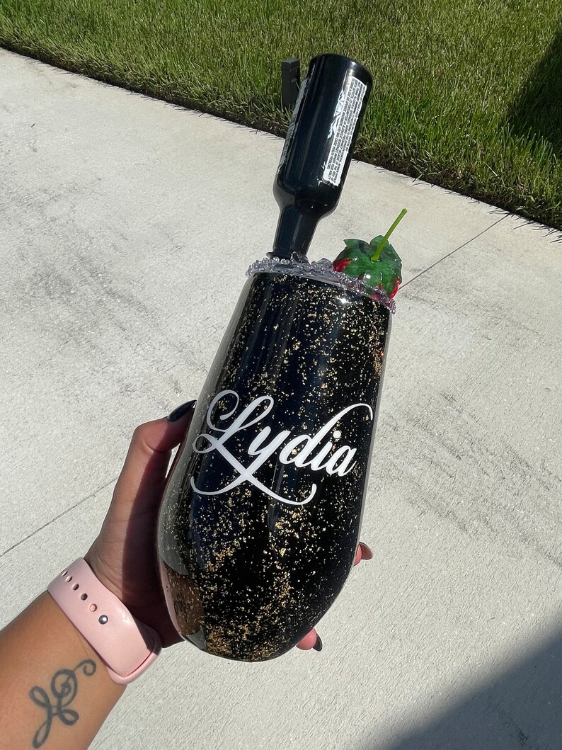 May include: Black and gold glitter tumbler with the name "Lydia" written in white cursive. The tumbler has a straw and a red strawberry garnish.
