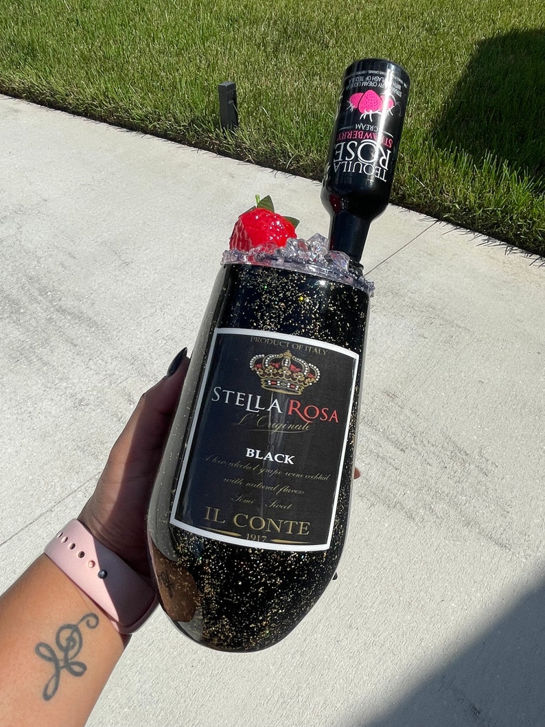 May include: A black and gold glitter tumbler with a Stella Rosa L'Originale Black label. The tumbler is topped with a red strawberry and a bottle of Stella Rosa Strawberry Cream Tequila.