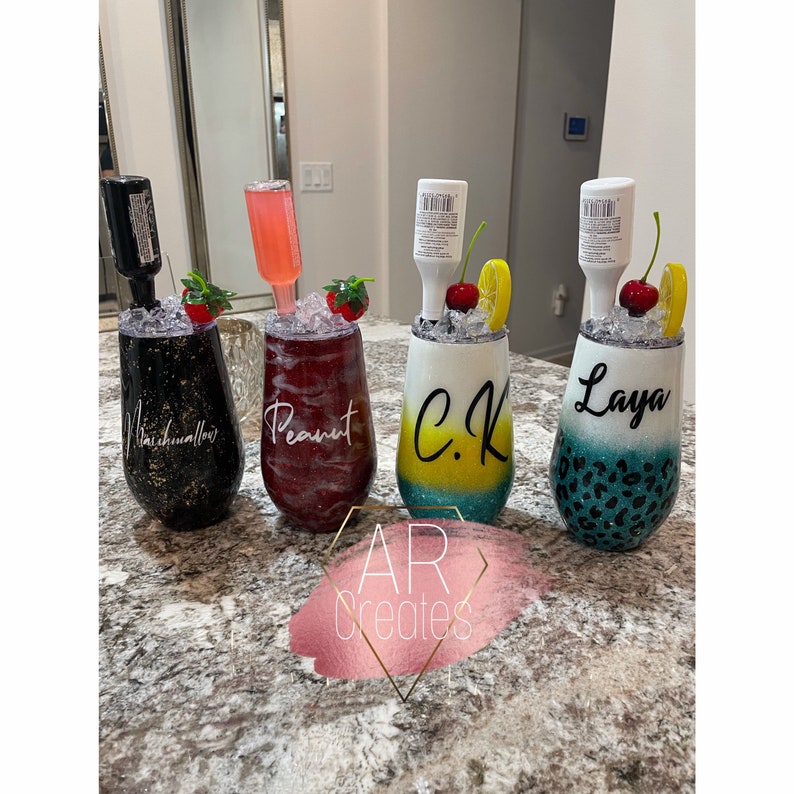 May include: Four personalized wine tumblers with different designs and colors. The tumblers are decorated with glitter, and each has a small bottle of liquor attached to the top. The tumblers are labeled "Marshmallow", "Peanut", "C.K." and "Layla".
