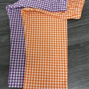 May include: Two pairs of checkered leggings, one purple and one orange. The leggings are folded and laid out on a dark wood surface.