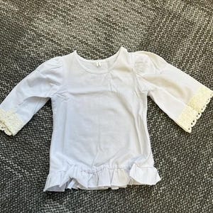 May include: A white long-sleeved shirt with a round neckline and ruffled hem. The sleeves have a bell shape with cream-colored lace trim at the cuffs. The shirt is made of a lightweight fabric.