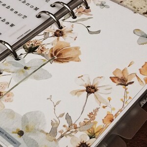 May include: A white planner with a floral design featuring watercolor flowers in shades of brown, beige, and light blue. The planner has a silver ring binding and the text "Lionheart Creations" is printed on the bottom left corner.