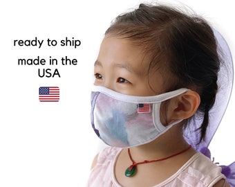 May include: A young person wearing a white face mask with a colorful abstract design. The mask has a white elastic band and a small American flag on the side. The person is wearing a pink sleeveless shirt and a green necklace.
