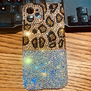 Może przedstawiać: Etui na telefon z wzorem lamparta pokryte kryształkami. Górna część etui to brązowo-czarny wzór lamparta, a dolna część jest w jasnoniebieskim, opalizującym kolorze.