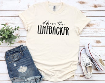Könnte beinhalten: Ein weißes T-Shirt mit dem Text "dibs on the LINEBACKER" in schwarzen Buchstaben. Das Shirt liegt auf einer weißen Holzoberfläche mit einer blauen Jeans-Shorts und weißen Turnschuhen.
