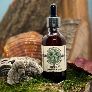 May include: A brown glass bottle with a black dropper, labeled "Emerald Coast Wellness Shoppe FORTIFY Turkey Tail & Reishi 1 fl oz / 30 ml." The bottle sits on a bed of green moss, with wood and fungi in the background.