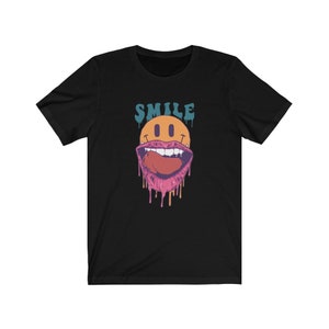 May include: Black t-shirt featuring a graphic of a smiling face with a large, open mouth and dripping pink saliva. The word "SMILE" is above the face in blue and orange lettering.