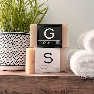 May include: Two bars of soap with black and white labels. The top bar has a black label with a white letter G and the text "Ganga Est 2008". The bottom bar has a black label with a white letter S and the text "Scherbaum Est 2008".