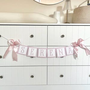 May include: A white dresser with six drawers, adorned with a banner spelling "SERENA" in pink letters. The banner is held by pink ribbons. A changing pad, lamp, and woven basket sit on top, suggesting a nursery theme.