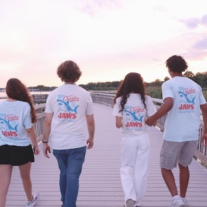 May include: Light blue short-sleeve t-shirts featuring a graphic on the back with a shark illustration, the text Charles, and the word JAWS in red lettering, worn by four individuals walking on a wooden pier during a sunset.