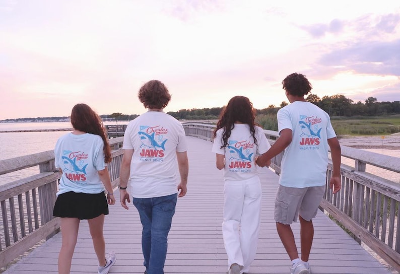 May include: Light blue short-sleeve t-shirts featuring a graphic on the back with a shark illustration, the text Charles, and the word JAWS in red lettering, worn by four individuals walking on a wooden pier during a sunset.