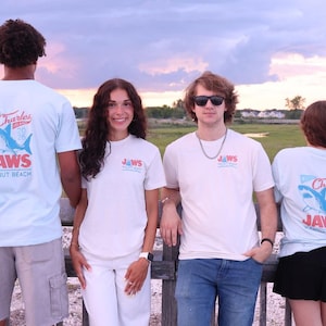 May include: Light blue and off-white t-shirts featuring a graphic on the back and front chest. The back design displays a blue shark illustration with the text Charles Island 2025 JAWS Walnut Beach. The front chest features a smaller matching logo.