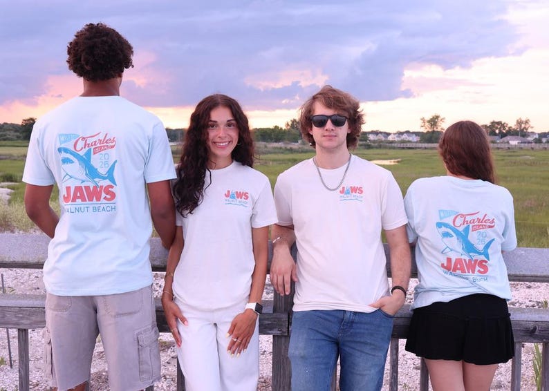 May include: Light blue and off-white t-shirts featuring a graphic on the back and front chest. The back design displays a blue shark illustration with the text Charles Island 2025 JAWS Walnut Beach. The front chest features a smaller matching logo.
