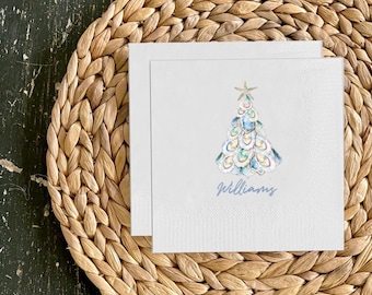 May include: Two white square napkins with a watercolor Christmas tree design. The tree is decorated with blue, green, and gold ornaments and topped with a starfish. The name "Williams" is written in blue script below the tree. The napkins rest on a woven placemat.