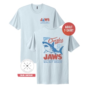 May include: A light blue short-sleeve t-shirt featuring a graphic of a shark. The front chest displays the text JAWS WALNUT BEACH. The back features a large shark illustration with the text MILFORD CHARLES ISLAND 2026 JAWS WALNUT BEACH.