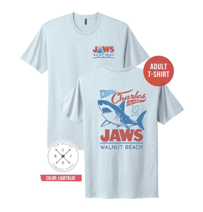 May include: A light blue short-sleeve t-shirt featuring a graphic of a shark. The front chest displays the text JAWS WALNUT BEACH. The back features a large shark illustration with the text MILFORD CHARLES ISLAND 2026 JAWS WALNUT BEACH.