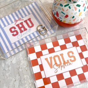 May include: Two clear acrylic trays with personalized designs. One features blue and white stripes with "SHU Charlotte" in red. The other has a red and white checkerboard pattern with "VOLS Sophia" in orange. Gold hoop earrings and a decorative vase are also present.