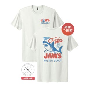 May include: A bone-colored short-sleeve t-shirt featuring a small graphic on the front chest with a shark and the text JAWS WALNUT BEACH. The back displays a large graphic of a shark with the text MILFORD CHARLES ISLAND 2026 JAWS WALNUT BEACH.