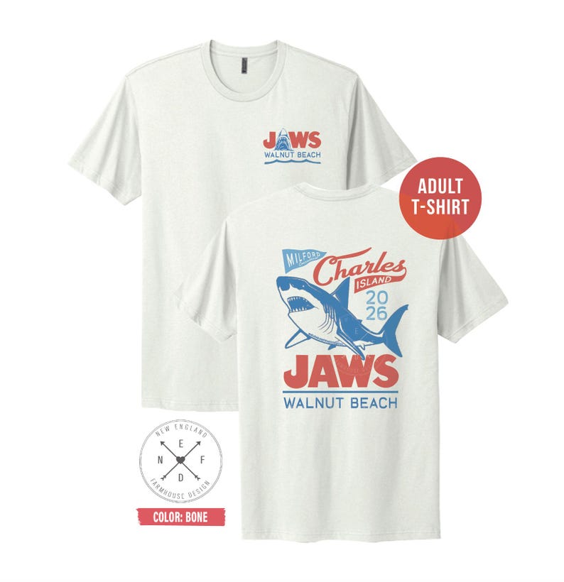 May include: A bone-colored short-sleeve t-shirt featuring a small graphic on the front chest with a shark and the text JAWS WALNUT BEACH. The back displays a large graphic of a shark with the text MILFORD CHARLES ISLAND 2026 JAWS WALNUT BEACH.