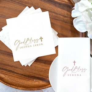 May include: White paper napkins with gold script text that reads "God Bless" and a cross. One stack of napkins has the name "Joshua Jakob" printed below the script. Another napkin has the name "Serena" printed in pink.