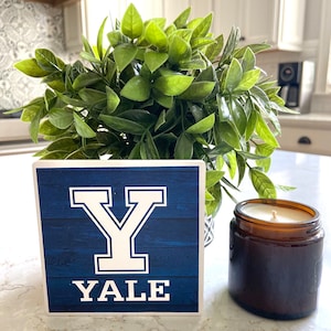 May include: A blue and white wooden sign with the letter "Y" and the word "YALE" in white lettering. The sign is sitting on a white countertop next to a brown glass candle.