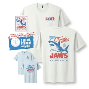 May include: Off-white and light blue t-shirts featuring a graphic of a shark and the text "Charles Island 2026 JAWS Walnut Beach". A matching sticker displays the same shark design and text. A blue rectangular sign reads "Limited Edition Merch".