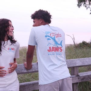 May include: A light blue short-sleeved t-shirt featuring a graphic on the back with a blue shark illustration, the text Charles Island 2025, the word JAWS in large red letters, and the text WALNUT BEACH printed in blue below the shark graphic.