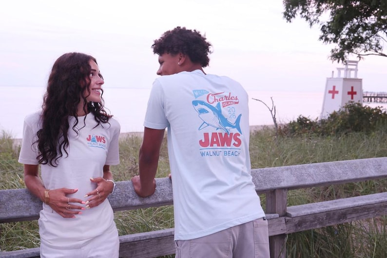 May include: A light blue short-sleeved t-shirt featuring a graphic on the back with a blue shark illustration, the text Charles Island 2025, the word JAWS in large red letters, and the text WALNUT BEACH printed in blue below the shark graphic.