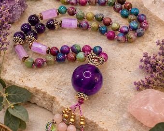 May include: A long beaded necklace featuring multicolored round stones in shades of pink, green, and blue, accented with cylindrical light purple beads, dark purple round beads, and a large central dark purple sphere with a decorative tassel.