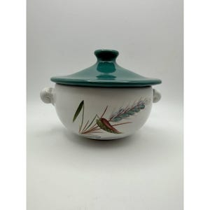 May include: A white ceramic soup bowl with a teal lid and handles. The bowl features a painted design of wheat stalks in green, red, and brown. The lid has a rounded knob. The bowl is suitable for serving soups and stews.