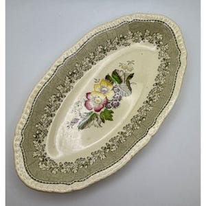 May include: An oval ceramic serving dish with a cream-colored center featuring a colorful floral bouquet of yellow, pink, and purple flowers. The wide border is decorated with a green oak leaf and acorn pattern on a textured background.