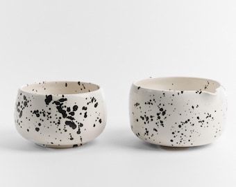 May include: Two white ceramic bowls with black splatter paint. The bowls are round and have a slight lip. The bowls are perfect for serving tea or other beverages.