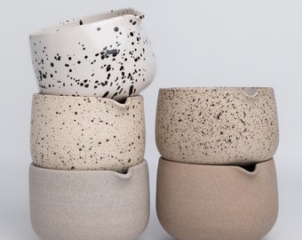 May include: A stack of five ceramic bowls in varying neutral tones. The top bowl is white with black speckles. The other bowls are beige and tan, with some speckled. Each bowl has a small spout.