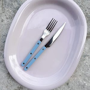 Puede incluir: Una fuente ovalada de color lavanda pálido con un tenedor y un cuchillo de acero inoxidable. El tenedor y el cuchillo tienen mangos azul claro con remaches negros. Los cubiertos están colocados en la fuente.