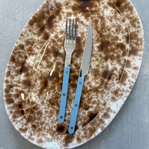 Op de afbeelding: Een ovale schaal met een bruin gespikkeld patroon, een vork en een mes. De vork en het mes hebben lichtblauwe handvatten. De set ligt op een grijze ondergrond.
