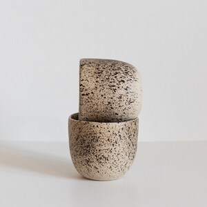 May include: Two beige ceramic mugs with a speckled brown glaze. The mugs are stacked on top of each other.
