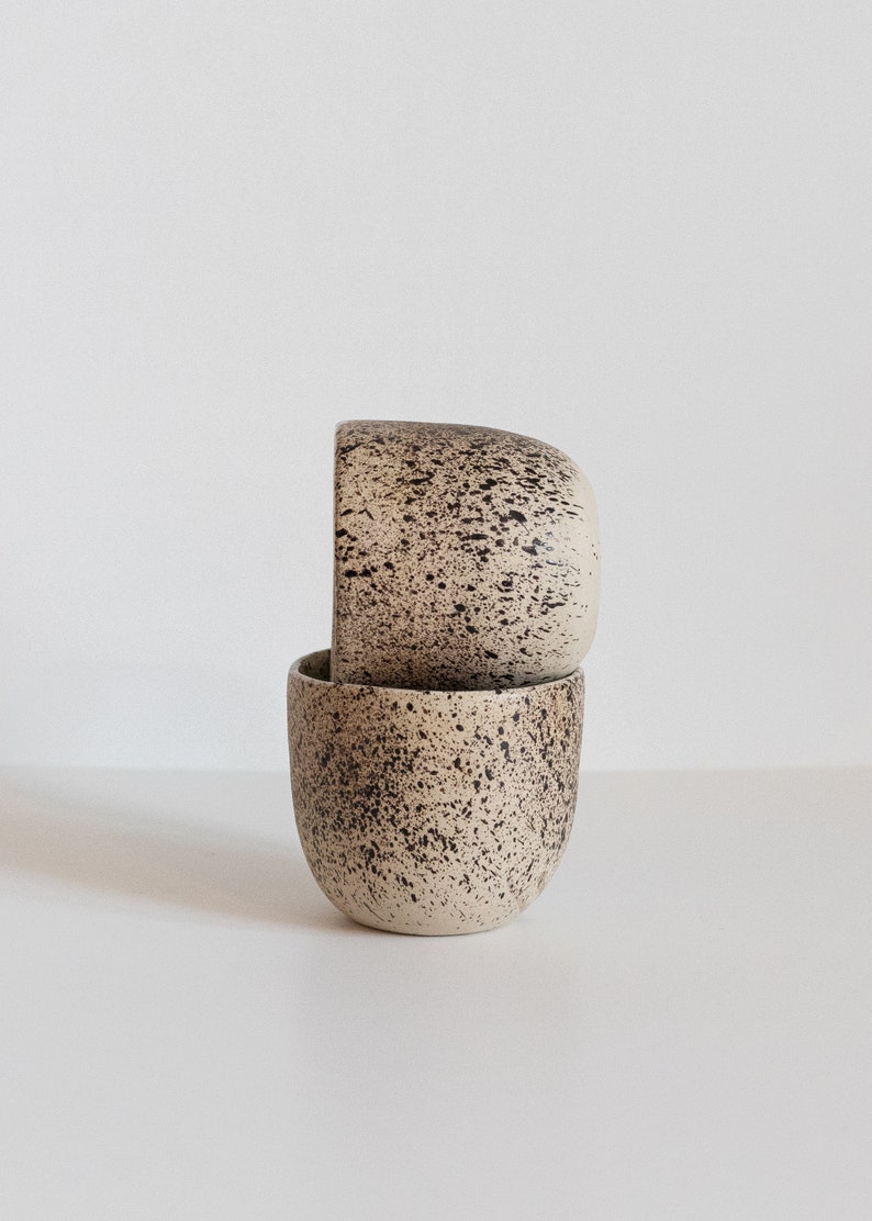 May include: Two beige ceramic mugs with a speckled brown glaze. The mugs are stacked on top of each other.