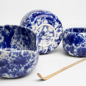 K&ouml;nnte beinhalten: Ein Set aus drei blauen und wei&szlig;en Keramik-Teetassen mit einem Spritzmuster. Die Tassen sind auf einer wei&szlig;en Oberfl&auml;che mit einem Bambus-Teebesen angeordnet.