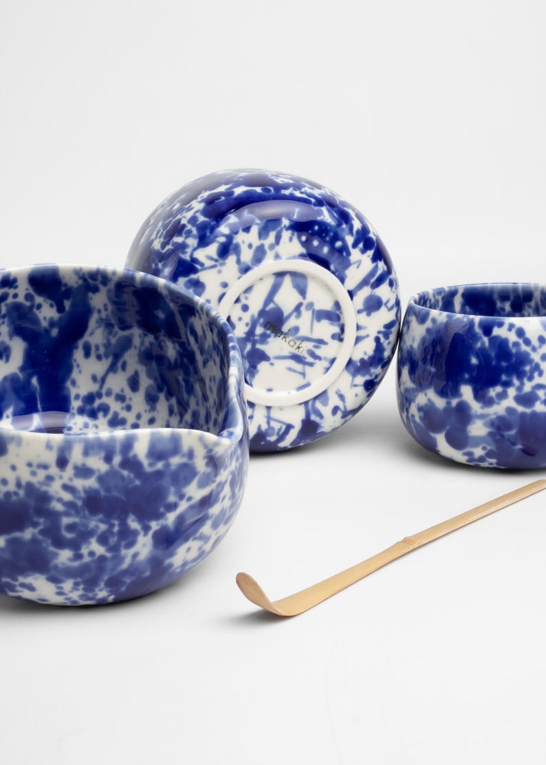 K&ouml;nnte beinhalten: Ein Set aus drei blauen und wei&szlig;en Keramik-Teetassen mit einem Spritzmuster. Die Tassen sind auf einer wei&szlig;en Oberfl&auml;che mit einem Bambus-Teebesen angeordnet.