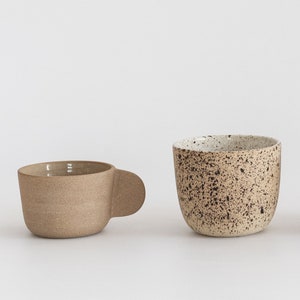 May include: Two ceramic mugs, one with a speckled brown and white glaze and the other with a solid brown glaze. Both mugs have a rounded shape and a handle.