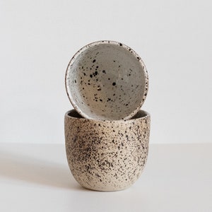 May include: Two ceramic mugs with a speckled brown and white glaze. The mugs are stacked on top of each other.