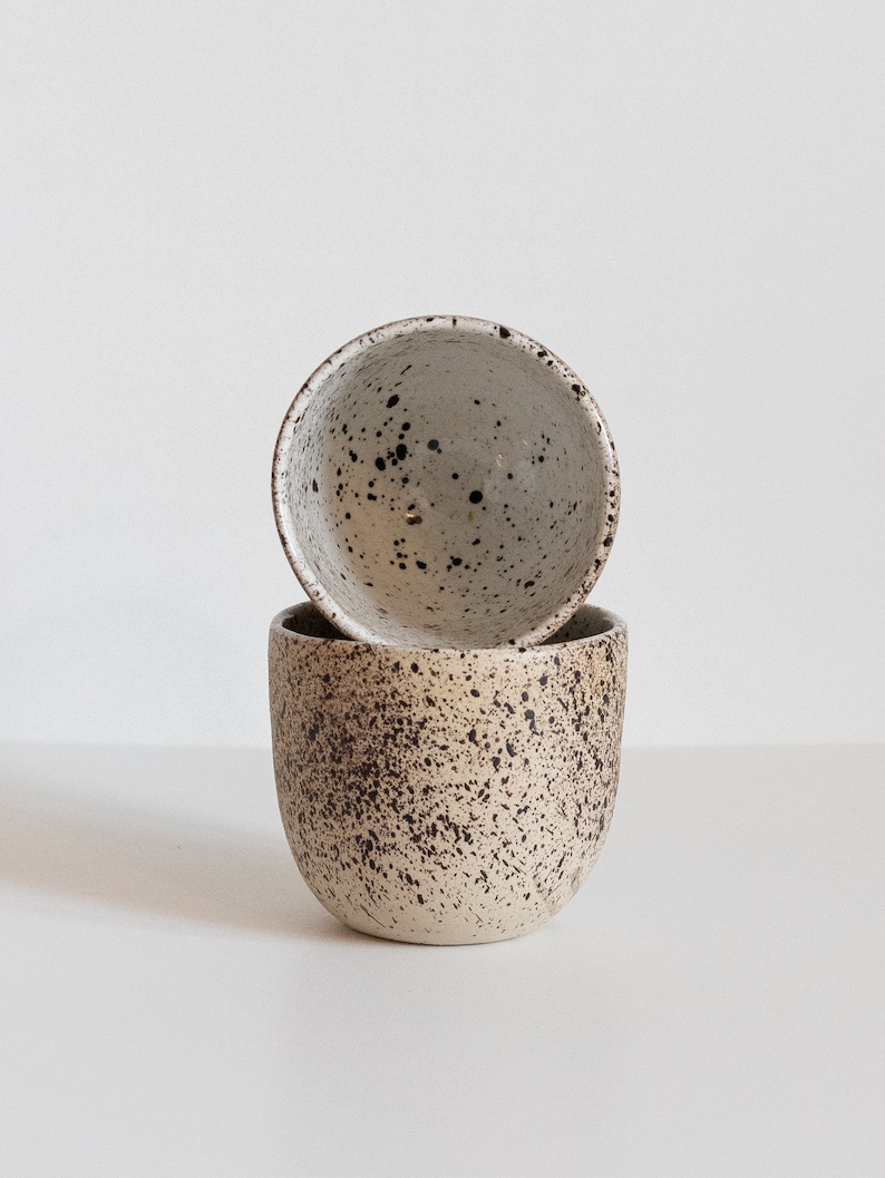 May include: Two ceramic mugs with a speckled brown and white glaze. The mugs are stacked on top of each other.