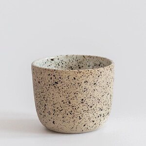 May include: A speckled ceramic cup with a light brown base and a white rim. The cup has a speckled pattern of black and white dots.