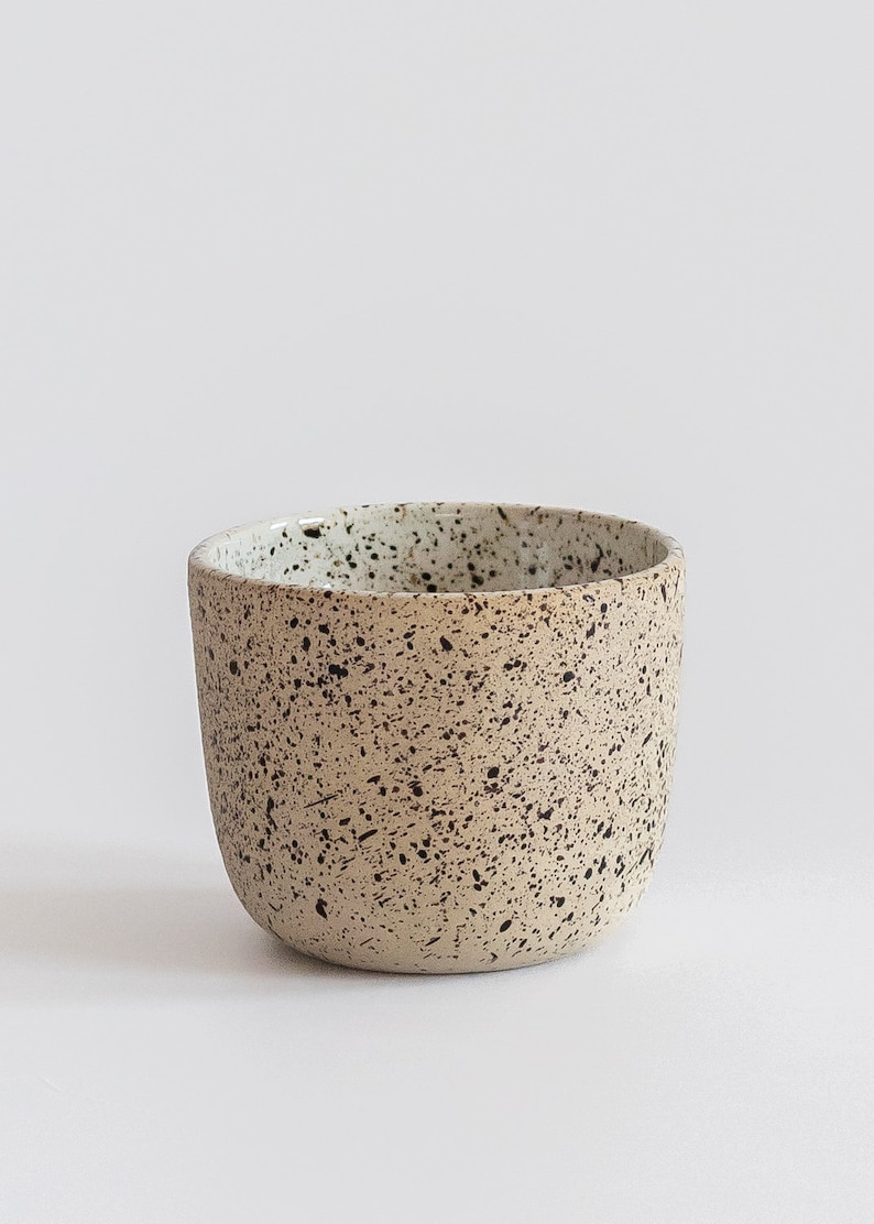 May include: A speckled ceramic cup with a light brown base and a white rim. The cup has a speckled pattern of black and white dots.