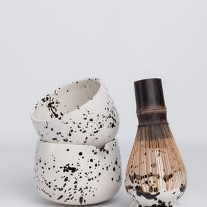 Puede incluir: Dos cuencos de cer&aacute;mica blanca con un dise&ntilde;o de pintura negra salpicada apilados uno encima del otro. Un batidor de matcha de bamb&uacute; con una base de cer&aacute;mica blanca y un dise&ntilde;o de pintura negra salpicada.