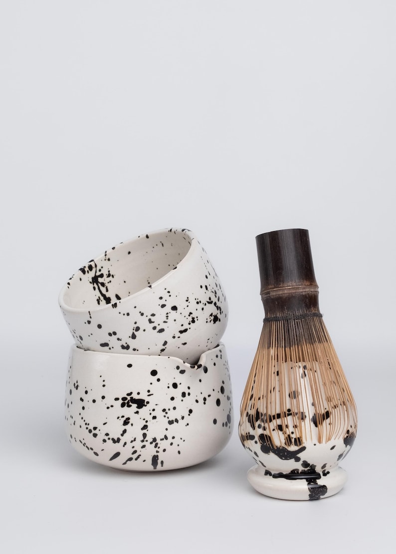 Puede incluir: Dos cuencos de cer&aacute;mica blanca con un dise&ntilde;o de pintura negra salpicada apilados uno encima del otro. Un batidor de matcha de bamb&uacute; con una base de cer&aacute;mica blanca y un dise&ntilde;o de pintura negra salpicada.