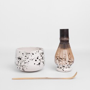 Puede incluir: Un cuenco de matcha de cer&aacute;mica blanca con un dise&ntilde;o de pintura salpicada negra, un batidor de matcha de bamb&uacute; con una base de cer&aacute;mica blanca con un dise&ntilde;o de pintura salpicada negra, y un batidor de matcha de bamb&uacute;.